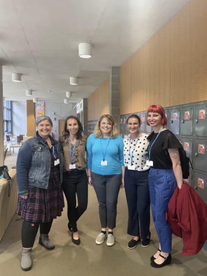 Five women stand in a hallway at a conference. They all have conference lanyards around their necks and are smiling at the camera.